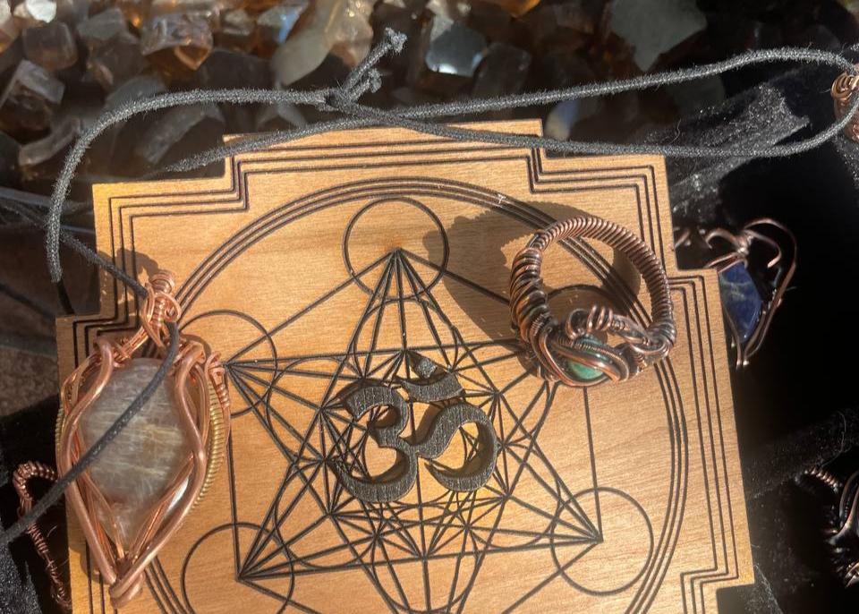 Wooden board engraved with a Metatron's Cube design and an Om symbol, surrounded by wire-wrapped jewelry and crystals in the background